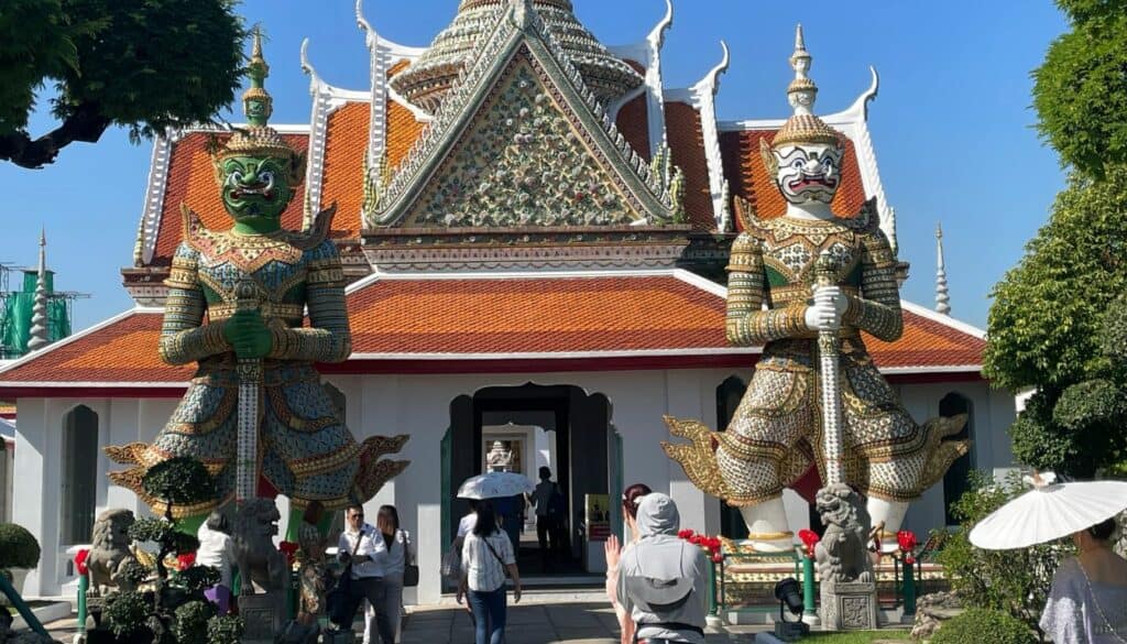 picture of Wat Arun temple in Bangkok Thailand. The picture shows a white temple with orange rooftop and has 2 guardian statues in front of it and tourists walking towards the house
