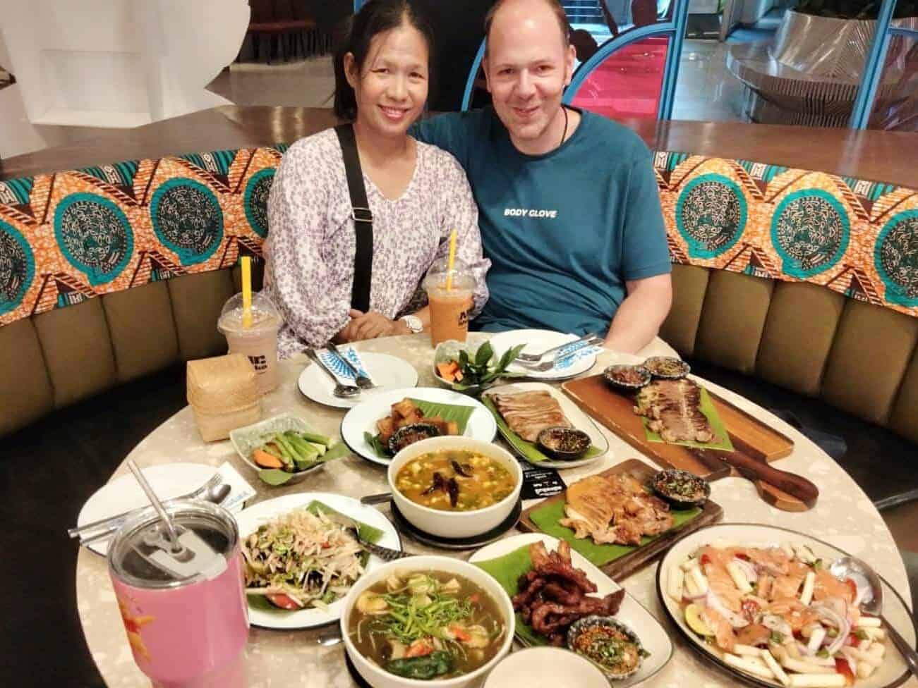in the image you'll see 2 people, Chris and Saengduan Verhoeven, from In Love With Thailand. They're sitting at a table in a restaurant and the table is filled with all kinds of different Thai food.