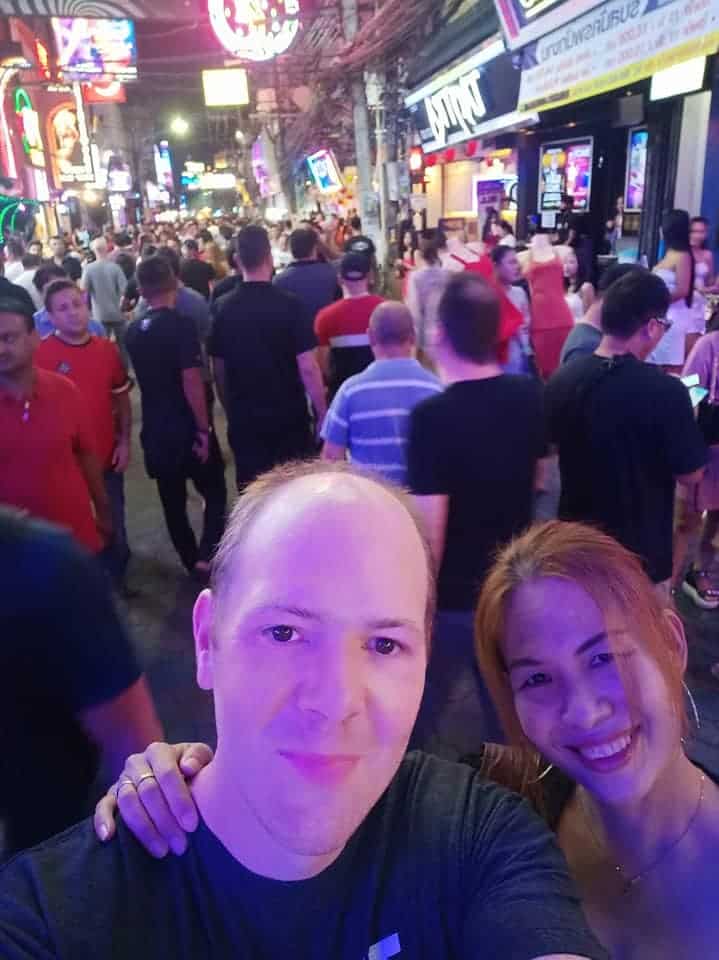the image shows a lot of people in Walking Street in Pattaya. At the bottom of the image it shows Chris and Saengduan Verhoeven from In Love With Thailand, looking into the camera. 

The people in the image are slightly blurred out and the image has many vibrant colors.