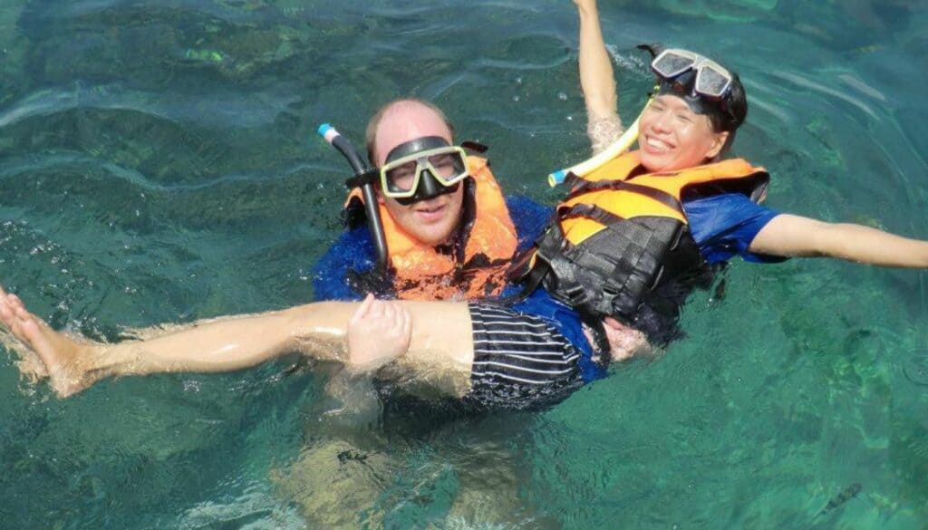 image of a man and a woman, Chris and Saengduan Verhoeven, who are snorkeling in Poda, which is a small island near Krabi. The image also shows turquoise water and Chris is holding Saengduan in his arms and both are wearing life vests and snorkeling gear.