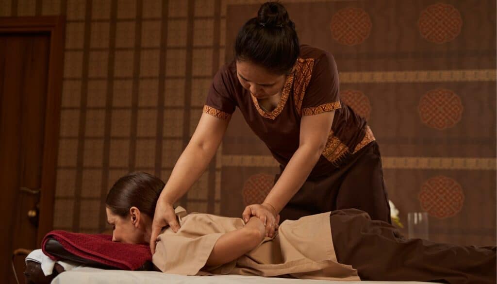 image of a woman getting a traditional Thai massage from a female Thai masseuse. The background is from wood and both women wear brown, neutral colored clothing.